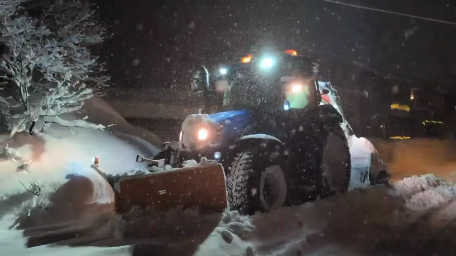 Blue snow-removal tractor clearing deep snow during a nighttime snowfall.